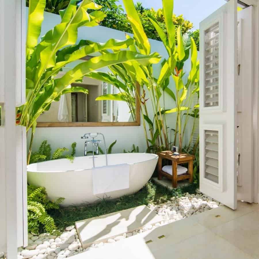 Outdoor bathroom with a white bathtub, surrounded by lush green plants, pebbles, and an open door leading to a sunny patio