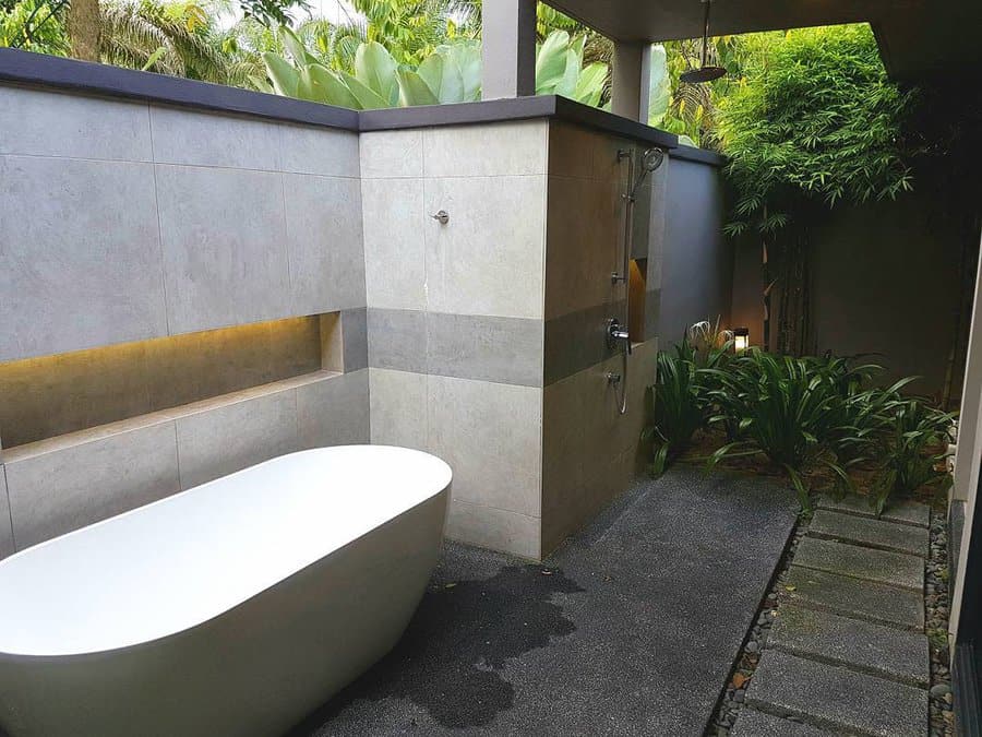 Outdoor bathroom with a freestanding bathtub, open shower, gray tiles, and lush greenery surrounding the space