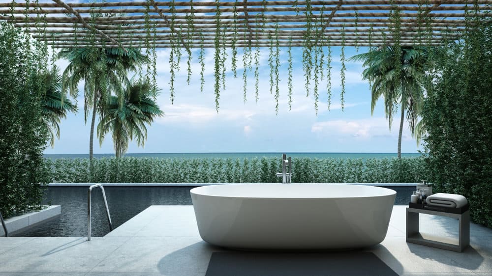 Outdoor bathtub overlooking the ocean, surrounded by greenery, palm trees, and a pergola with hanging vines