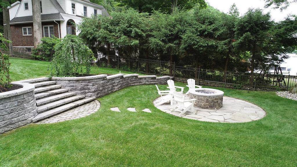 Beautiful backyard with a curved retaining wall, stone steps, circular fire pit area, and Adirondack chairs surrounded by lush greenery