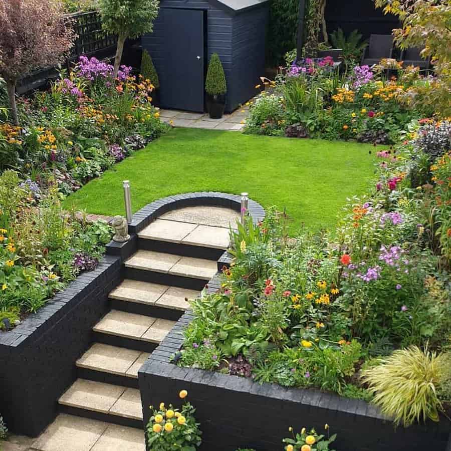 Vibrant backyard garden with colorful flower beds, a lush green lawn, black brick retaining walls, and stone steps leading to a storage shed