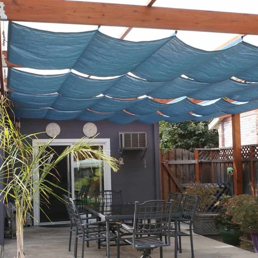 Outdoor patio with a wooden pergola featuring blue retractable shade panels, a black metal dining set, and lush greenery