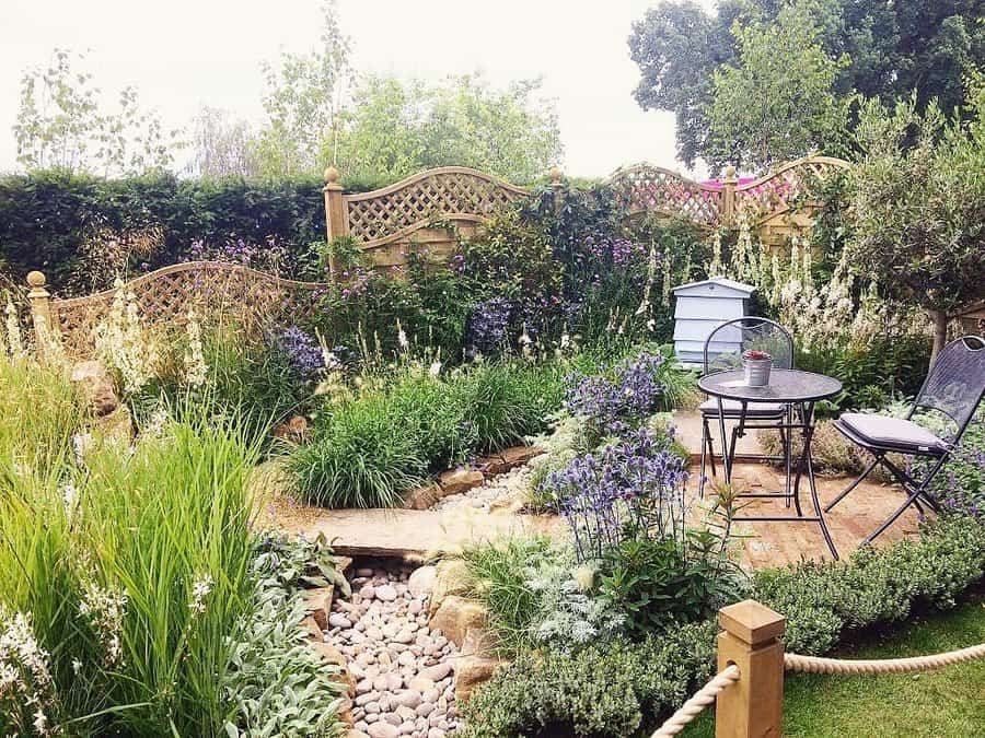 Charming garden retreat featuring a cozy bistro set on a stone patio, surrounded by lush greenery and vibrant flowers. A dry creek bed adds a natural touch, while wooden trellises create a private and tranquil ambiance, perfect for relaxation.