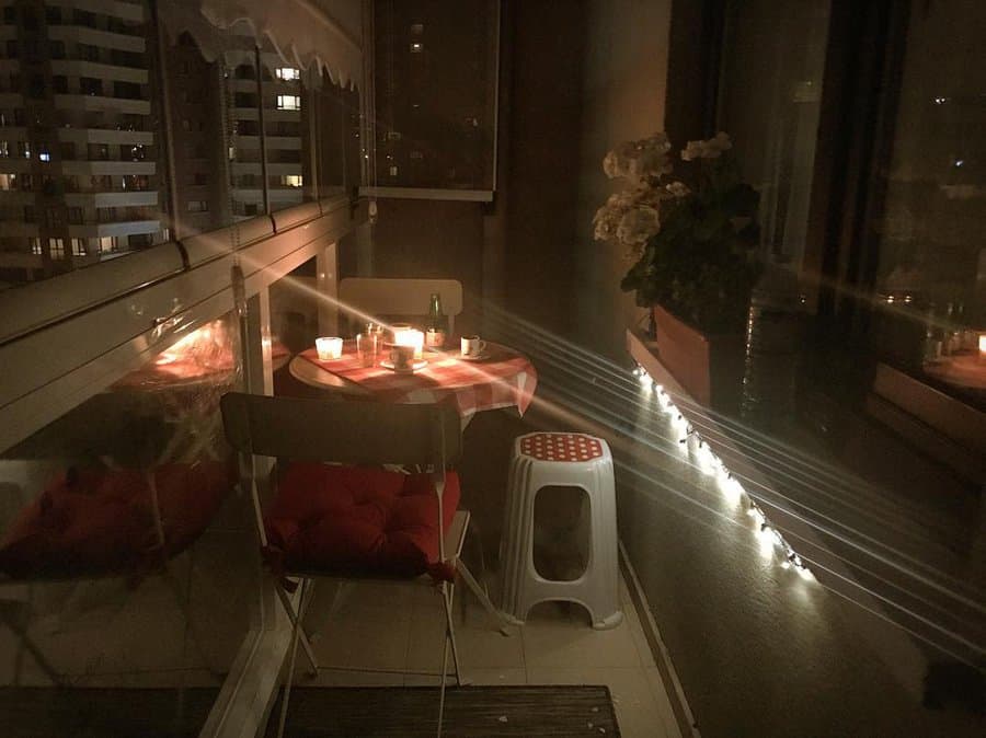 Romantic small balcony at night with a round table covered in a checkered cloth, candles, and a cozy chair with a red cushion. String lights and a potted plant add warmth to the intimate setting.