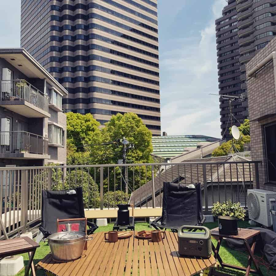 Urban rooftop balcony with a wooden table, camping chairs, and greenery, offering a cozy outdoor retreat with city skyline views