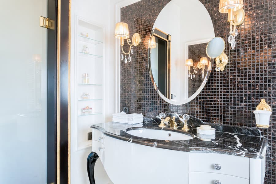 Elegant bathroom with dark mosaic tiles, oval mirror, gold-accented sconces, and a black marble countertop with a white sink