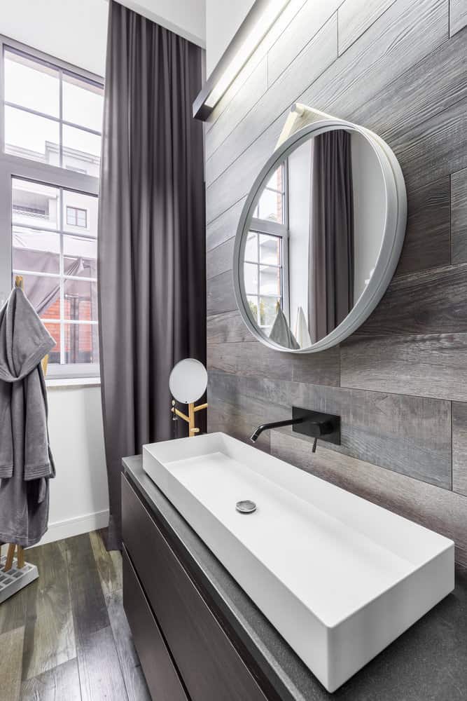 Modern bathroom with a rectangular white sink, round mirror, dark wood accents, black faucet, and a window with gray curtains