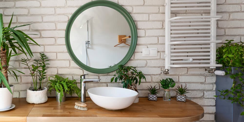 Bathroom with a round green mirror, white sink, wooden countertop, plants, and a white towel rack on a brick wall