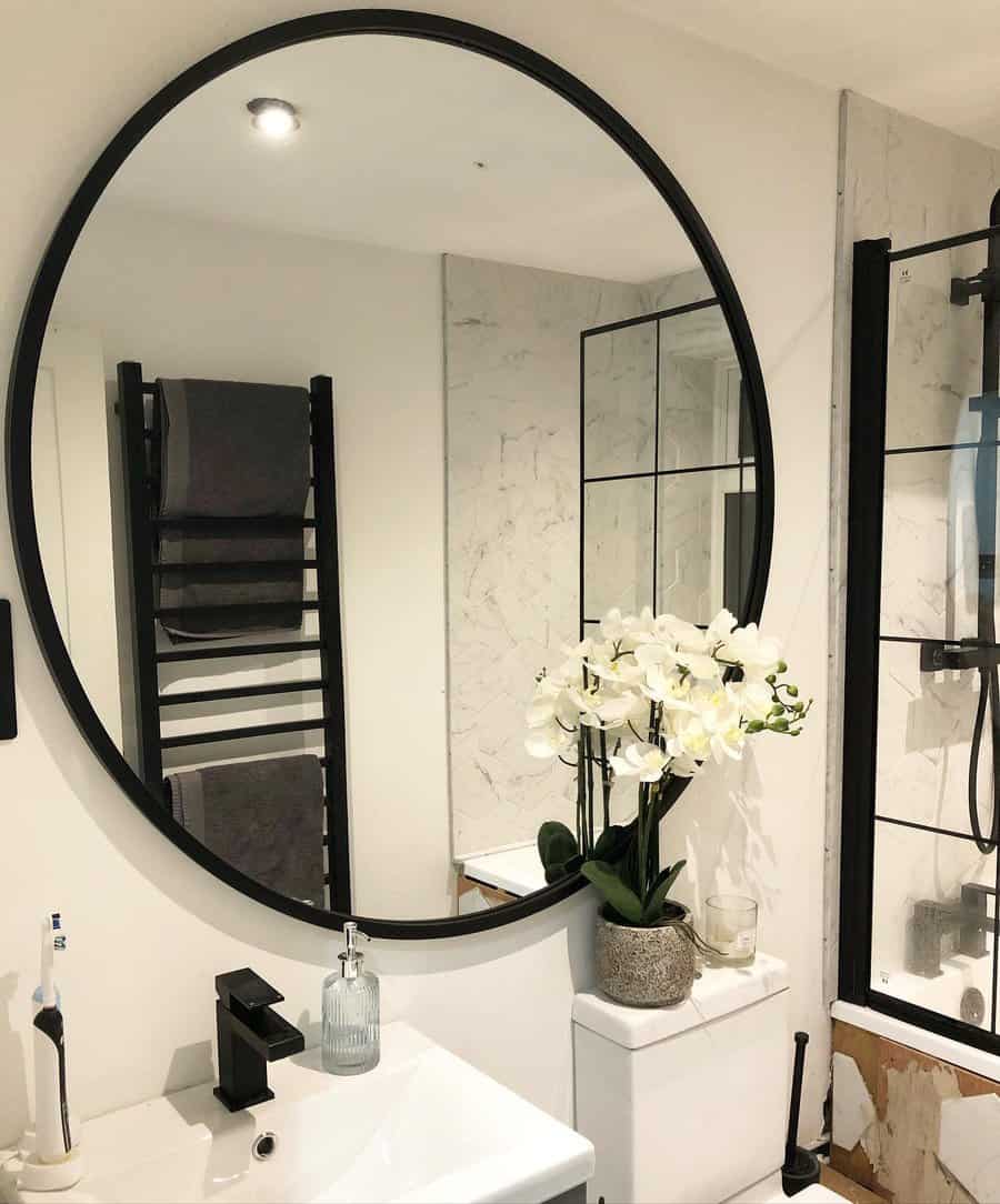 Modern bathroom with a large round mirror, white sink, black fixtures, a towel rack, and an orchid plant on a white toilet tank