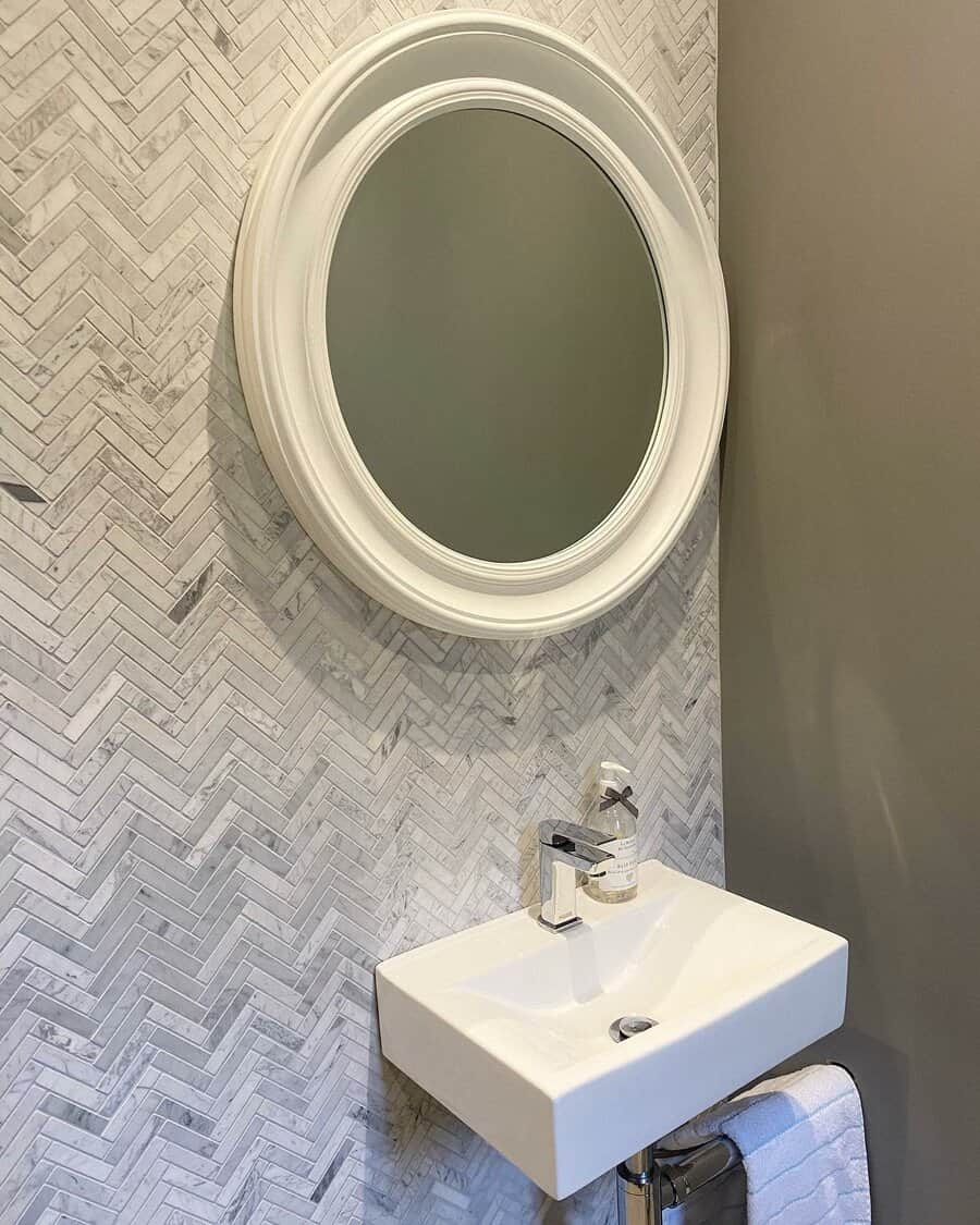 Small bathroom with a round mirror above a white square sink, herringbone patterned tile wall, and a hand towel beneath the sink