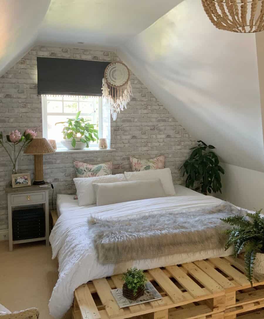 Cozy attic bedroom with a boho touch, featuring a pallet bed, soft textures, greenery, and a brick accent wall for a relaxed, rustic vibe.