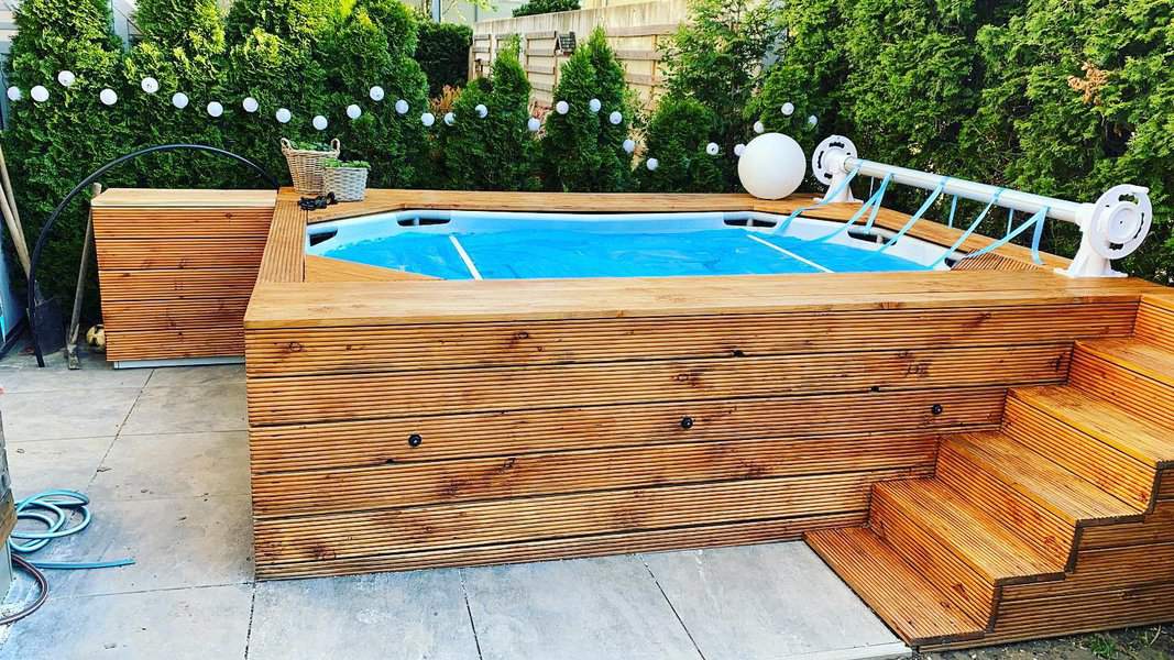 Wooden deck surrounds a small above-ground pool with blue water, steps lead up to the pool framed by green shrubs and decorative lights