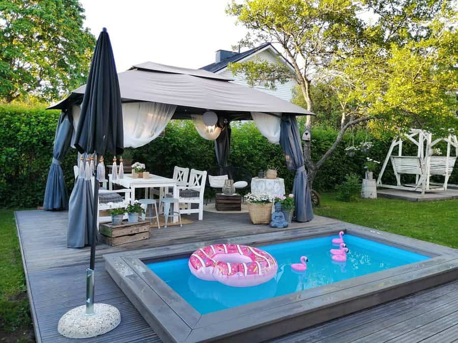 Backyard setup with a gazebo, dining table, small pool with inflatable donut and flamingos, surrounded by greenery and a swing