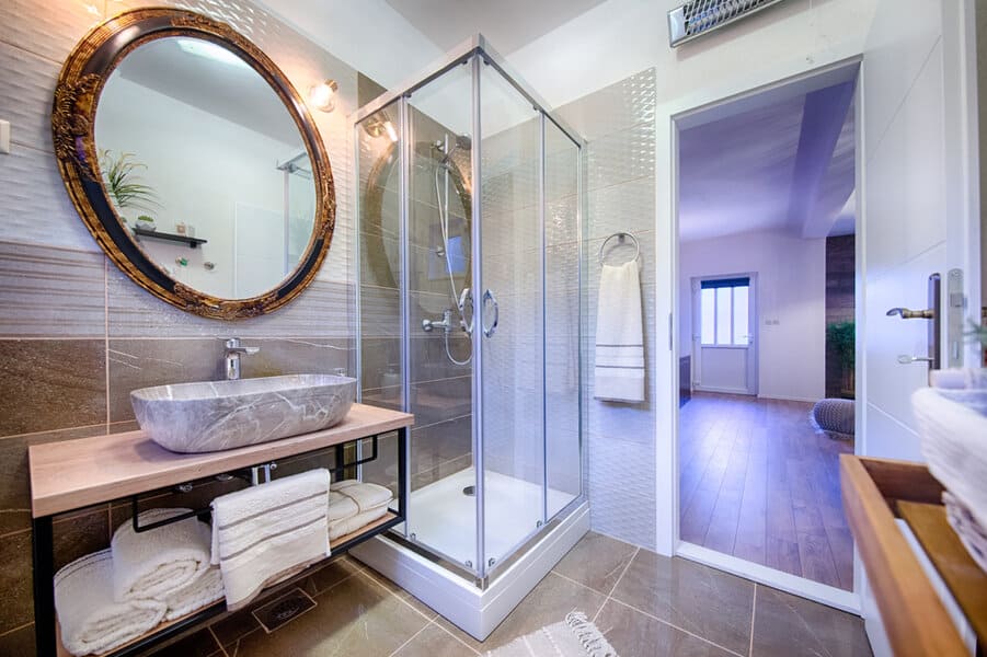 Modern bathroom with a glass shower, round mirror, stone sink, and wooden accents; open door leads to a bright room with wooden flooring