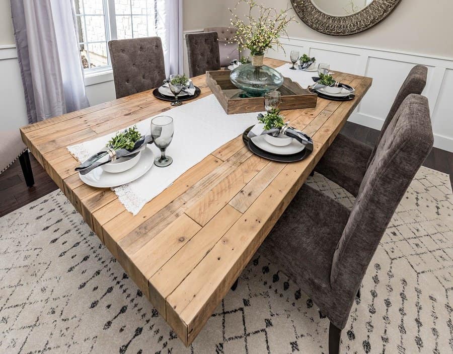 A rustic wooden dining table set with plates, glasses, and greenery, surrounded by four upholstered chairs on a patterned rug