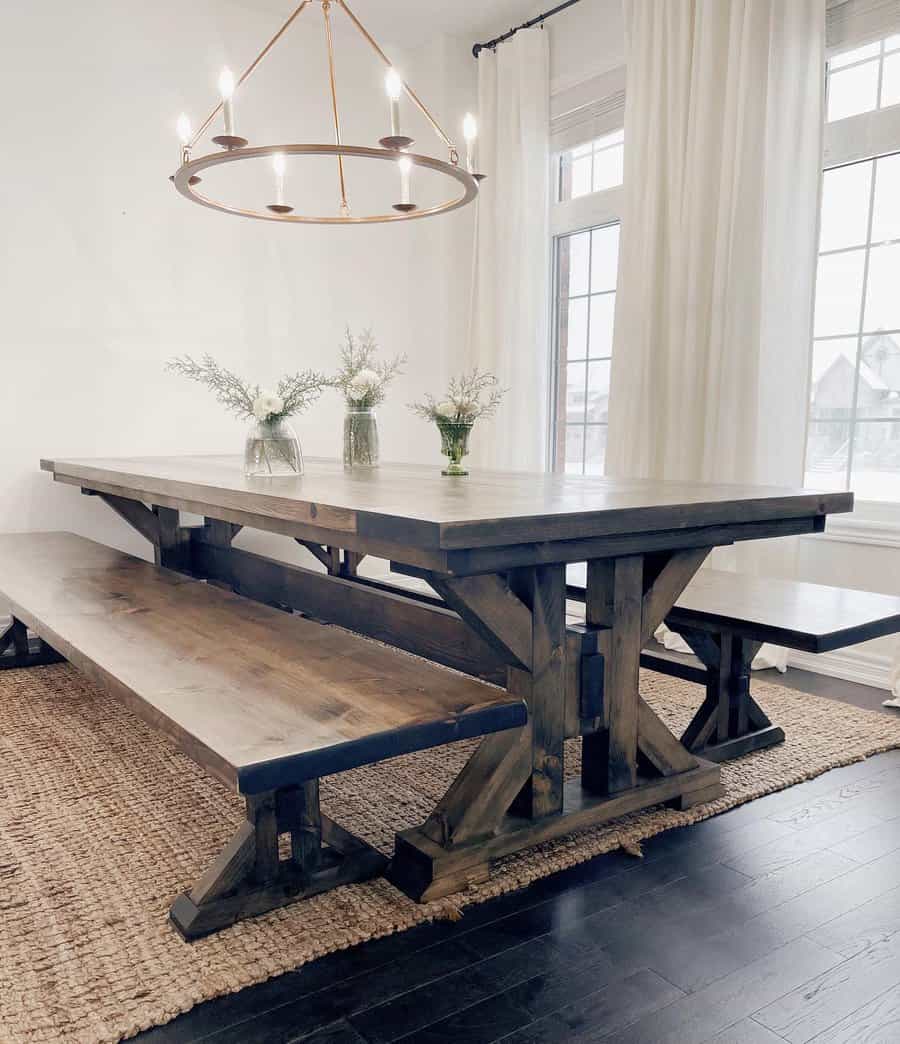 Rustic dining room with a large wooden table, matching benches, a round chandelier, and vases of flowers, set against white curtains