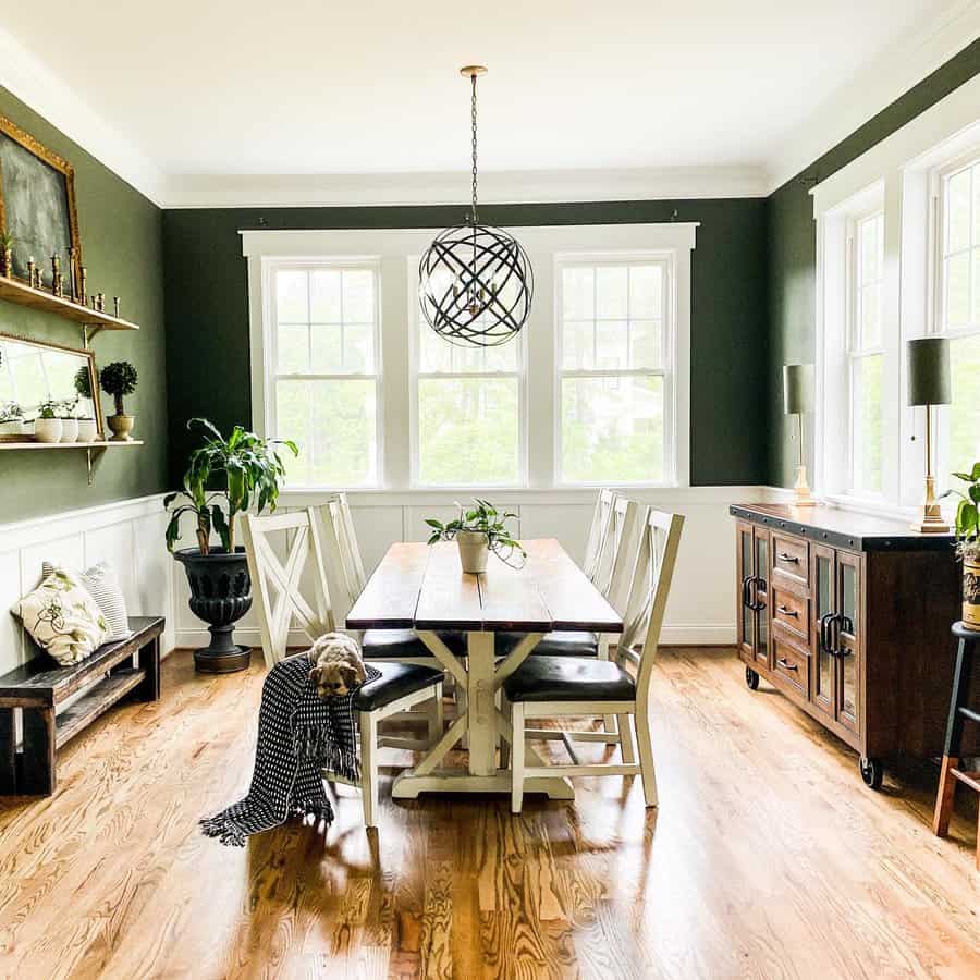 A farmhouse dining room with a wooden table, six chairs, potted plants, a hanging light fixture, and large windows with natural light