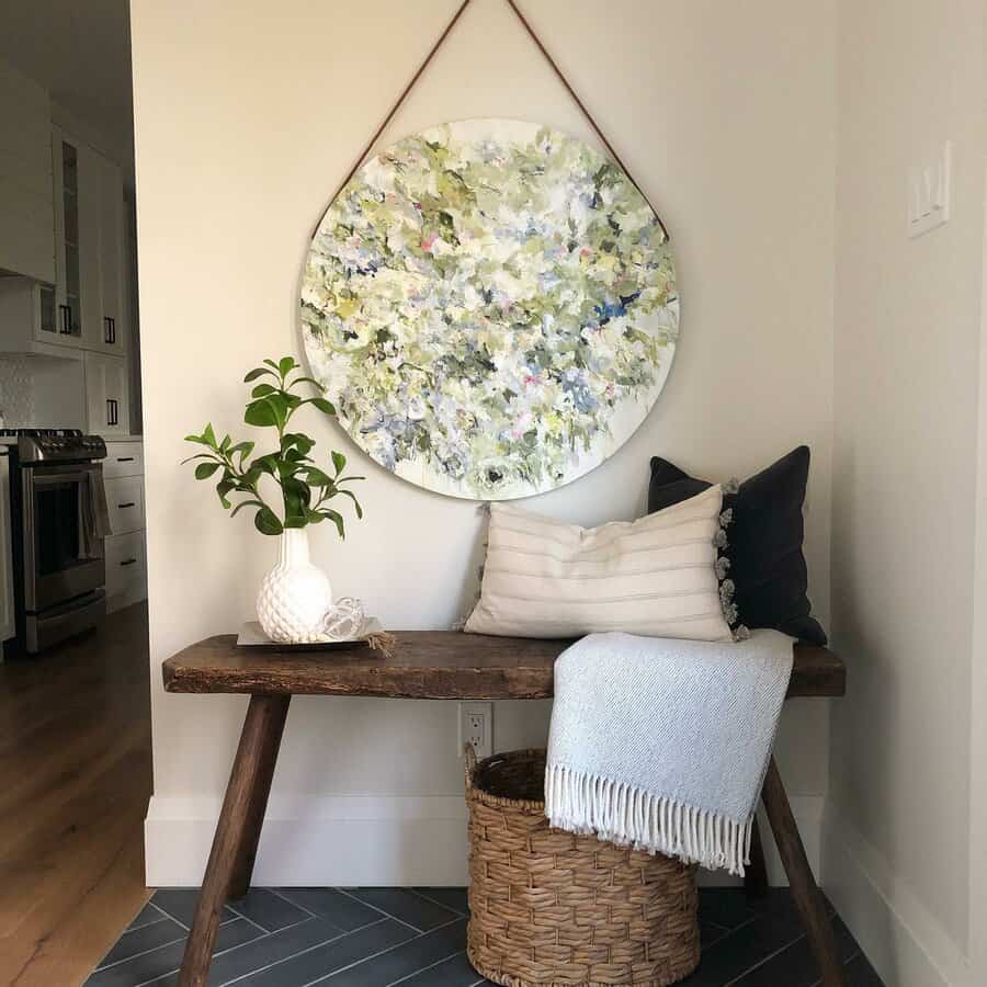 Wooden bench with pillows and a blanket, potted plant, wicker basket beneath, and abstract circle painting on wall in bright hallway