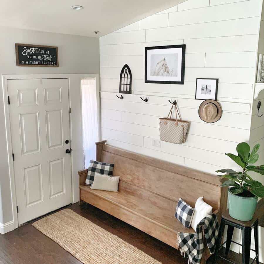 Cozy entryway with a wooden bench, plaid pillows, wall hooks with bags and hats, and framed photos; white shiplap wall and a plant