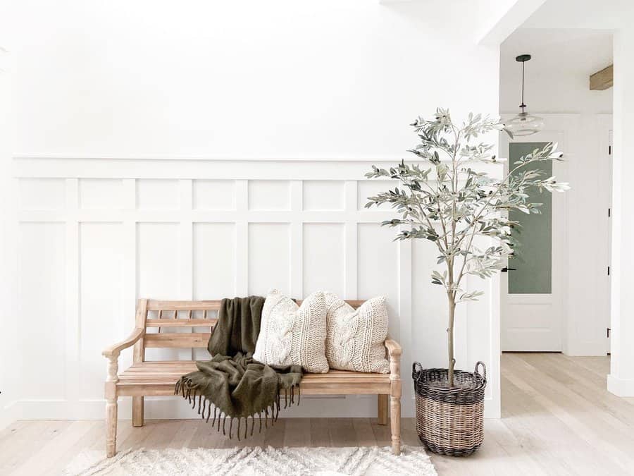 Minimalist entryway with a wooden bench, cozy pillows, green throw, and potted plant; white paneled wall and soft lighting