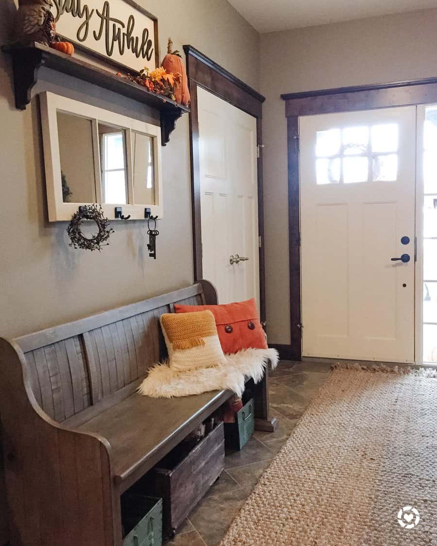 Cozy entryway with a wooden bench, pillows, and decor; mirror and sign on wall; light-colored door, natural rug, and small wreath