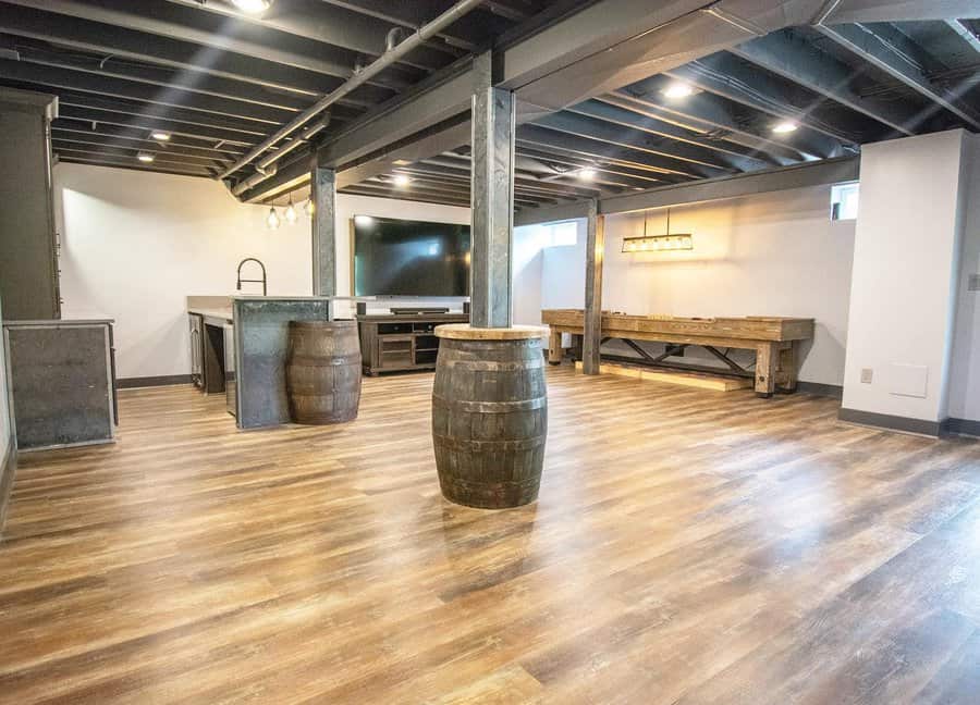 Spacious rustic basement with an open ceiling, wooden flooring, whiskey barrel accents, a built-in bar, and a shuffleboard table for entertainment.