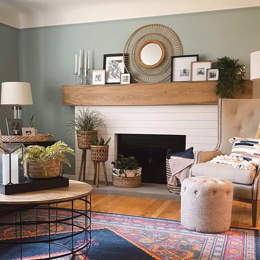 Cozy living room with a green accent wall, wooden mantel, plants, a round mirror, and a tufted chair; patterned rug and round coffee table