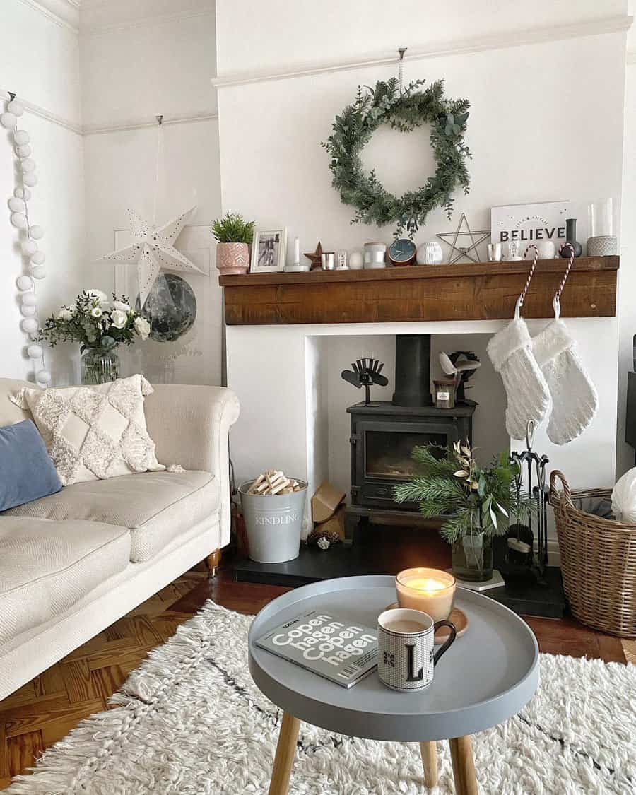 Cozy living room with a white sofa, wood fireplace, stockings, wreath, candles, and a small table with a candle and mug