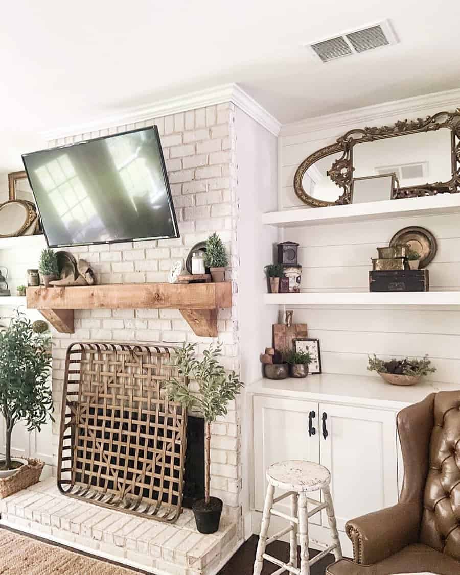 Cozy living room with a brick fireplace, TV, wooden mantel, mirrors, shelves with decor, and a brown leather chair