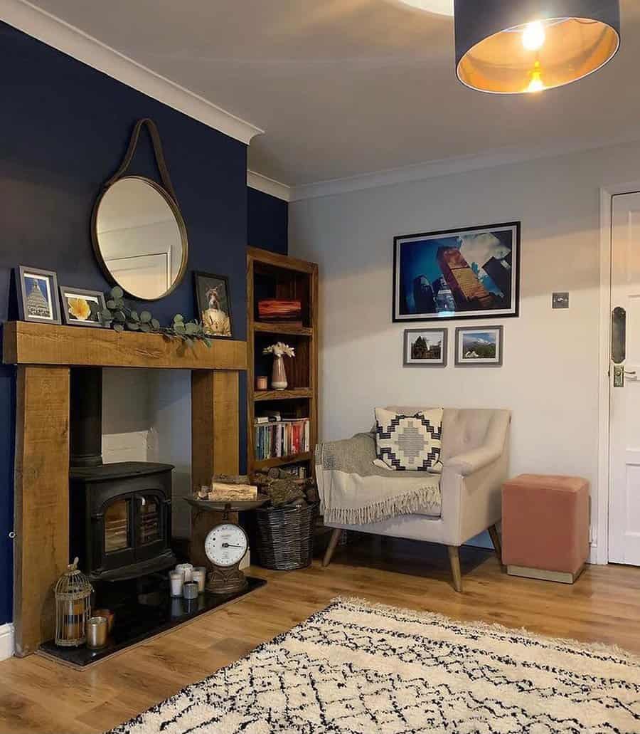 Cozy living room with a wood-burning stove, a round mirror, bookshelves, an armchair with a throw, and decorative items on the mantel