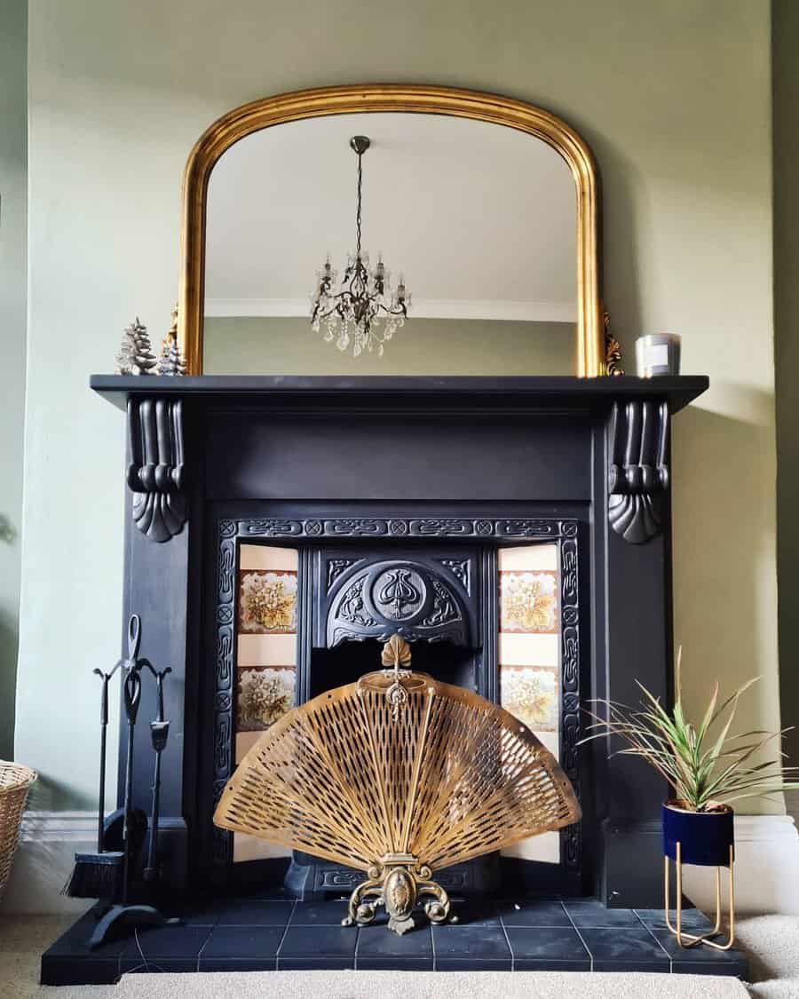 A classic black fireplace with a golden-framed mirror above, ornate detailing, and a decorative brass fan, complemented by vintage tiles and plants for a timeless look