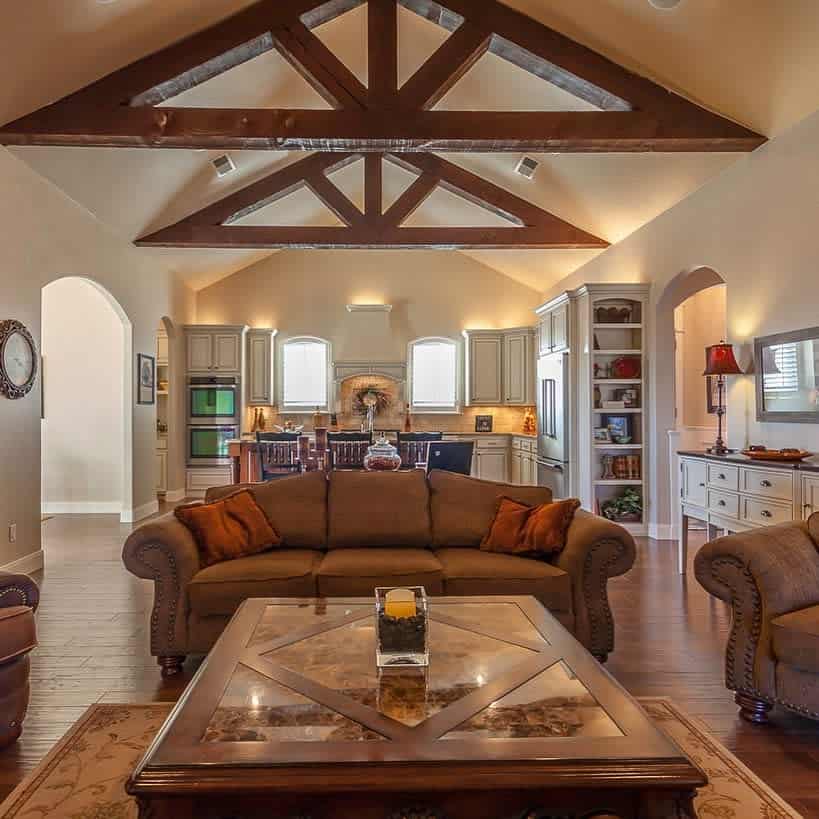 Spacious living room with exposed wooden beams, brown sofas, a glass coffee table, and a view into a cozy kitchen area