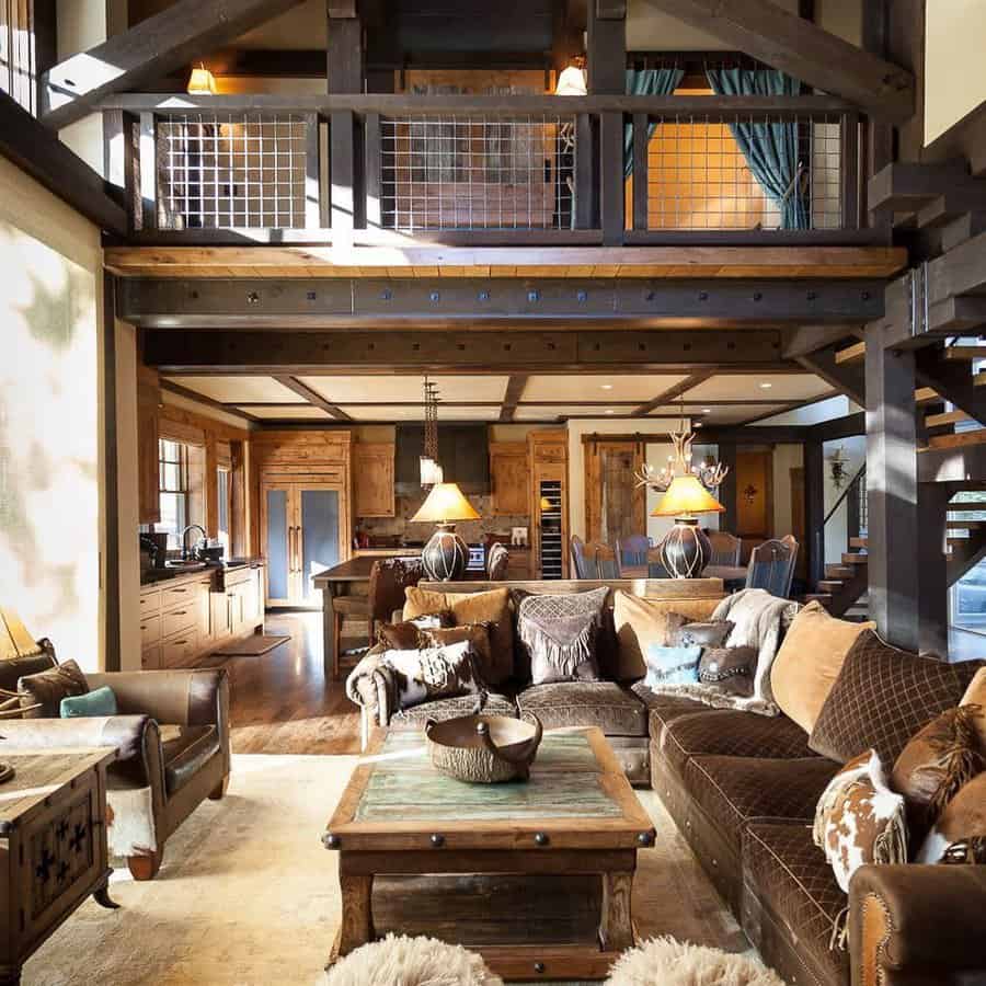 Rustic living room with exposed beams, brown leather couches, a wooden coffee table, and a loft area with a staircase