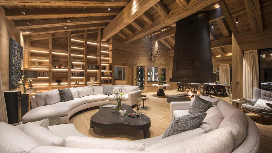 Spacious living room with curved sofas, wooden beams, built-in shelves, large fireplace, and a dining area in the background