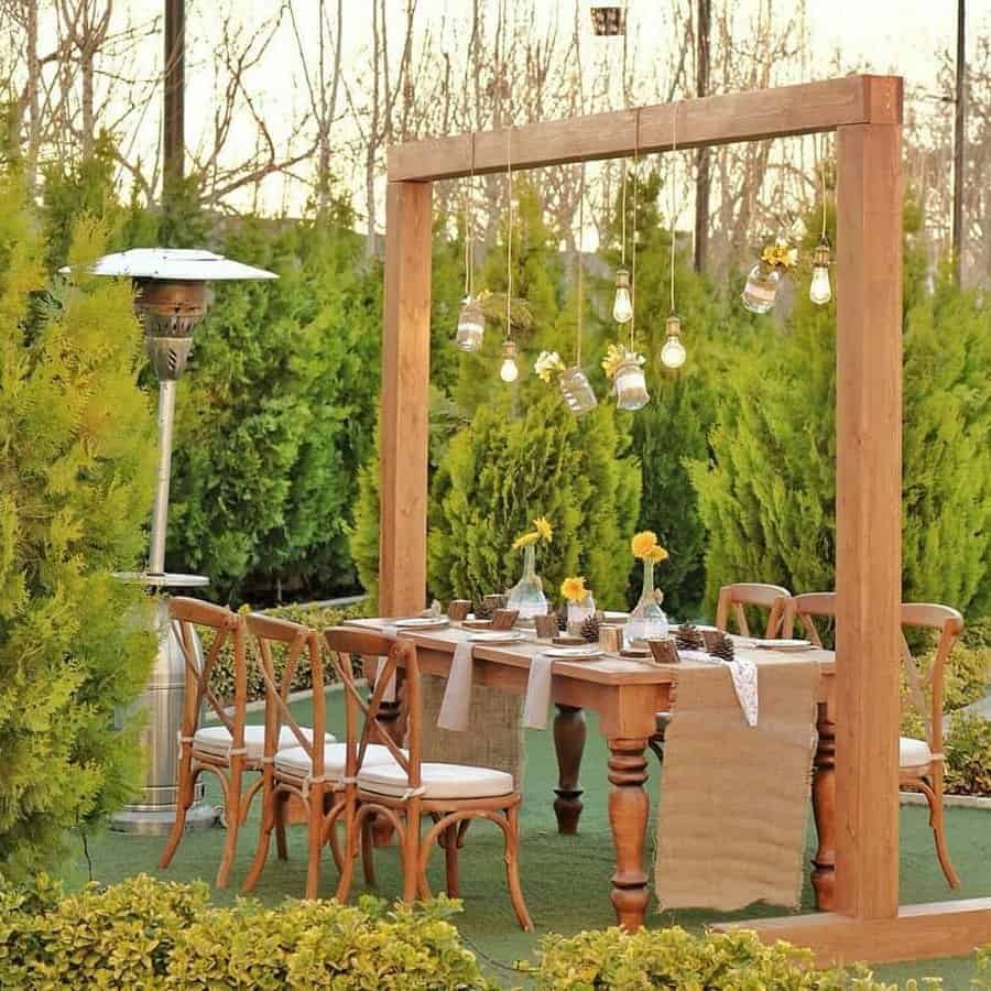 Outdoor dining setup with wooden table and chairs, surrounded by greenery, with hanging lights in jars above the table creating a cozy ambiance