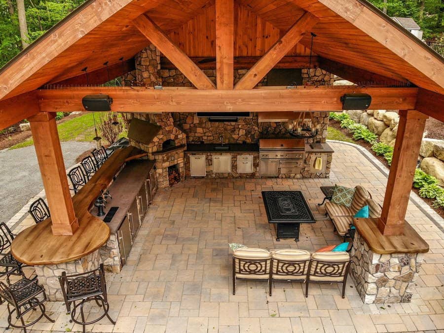 Outdoor kitchen with stone and wood design, featuring bar stools, grill, fireplace, and seating area under a wooden pavilion