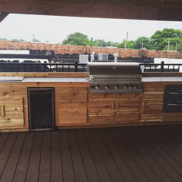 Outdoor kitchen with wooden cabinets, stainless steel grill, and mini fridge on a wooden deck; stone wall and greenery in the background