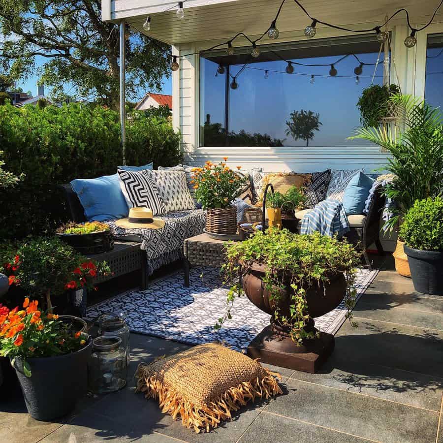 Cozy outdoor patio with cushioned seating, plants, string lights, and a woven rug, surrounded by greenery on a sunny day