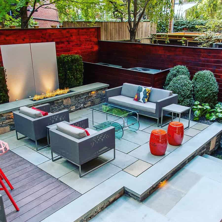 Modern outdoor patio with gray seating, a glass coffee table, red stools, and a stone fireplace surrounded by lush greenery