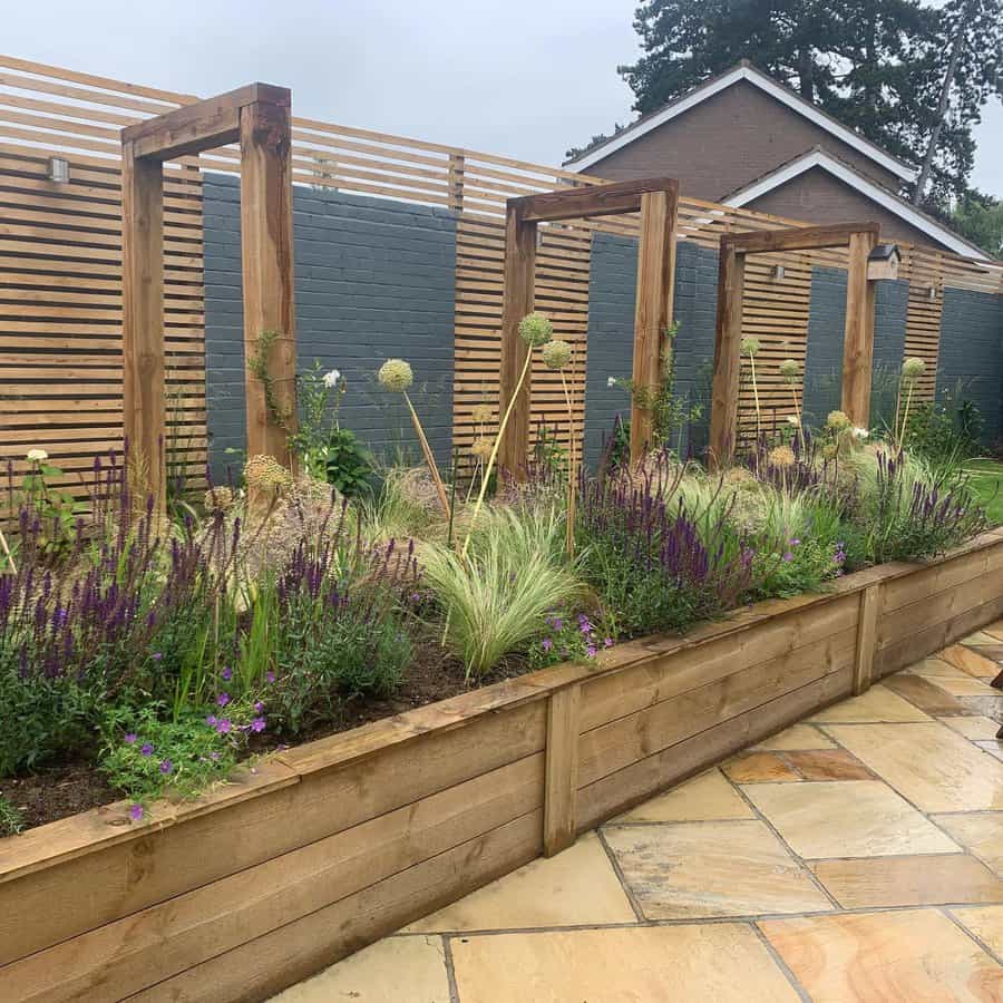 Modern garden with wooden pergolas, slatted fencing, raised flower beds, ornamental grasses, and purple blooms on a stone patio