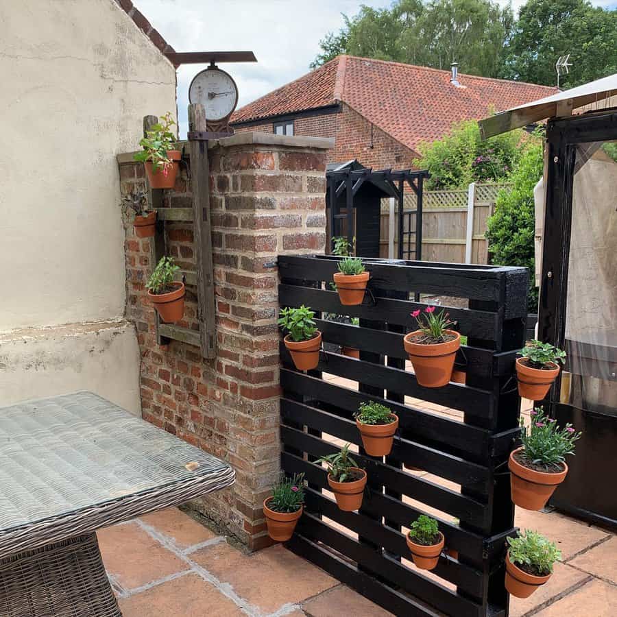 Outdoor patio with a black wooden pallet displaying potted plants, brick wall partially visible; a table is on the left side