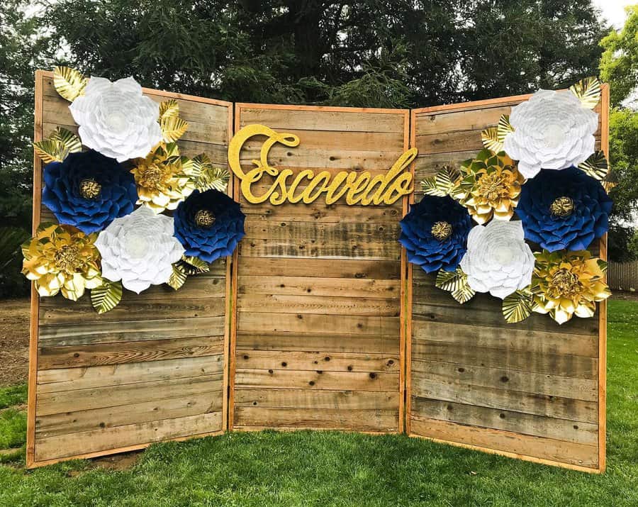 Wooden backdrop with large blue, white, and gold paper flowers and "Escovedo" written in gold cursive, lush trees in the background