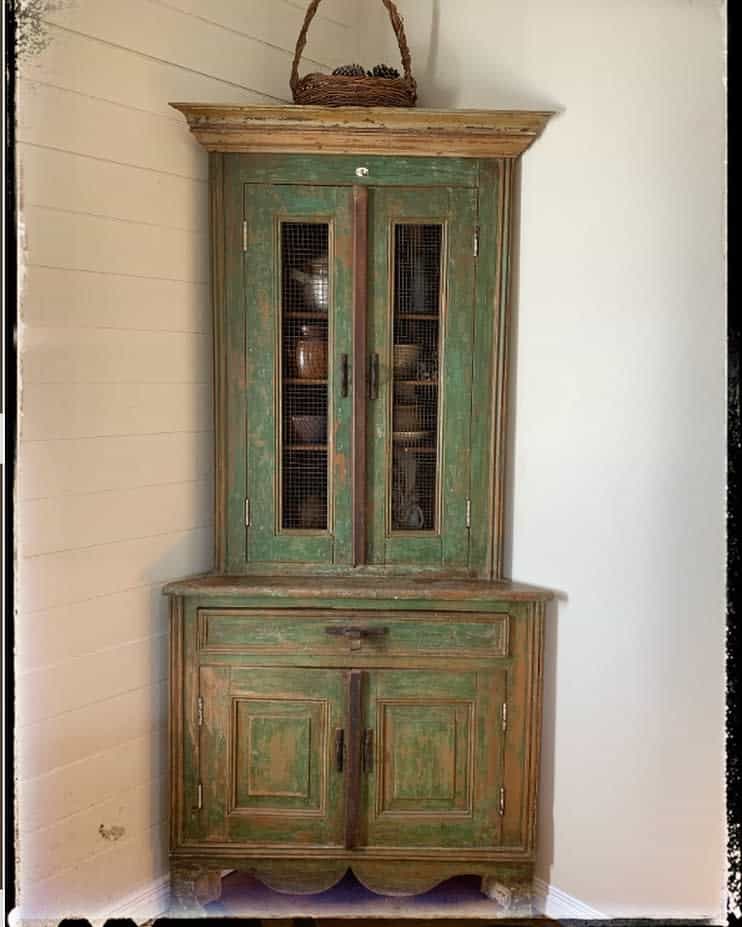 Rustic green distressed corner hutch with wire mesh doors and a weathered wood finish, styled with a woven basket on top.