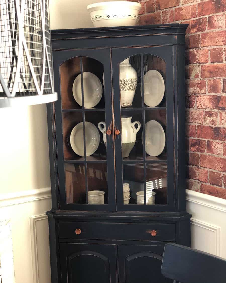 Vintage black-painted corner hutch with glass doors and a distressed finish, styled with white dishware against a rustic brick wall.