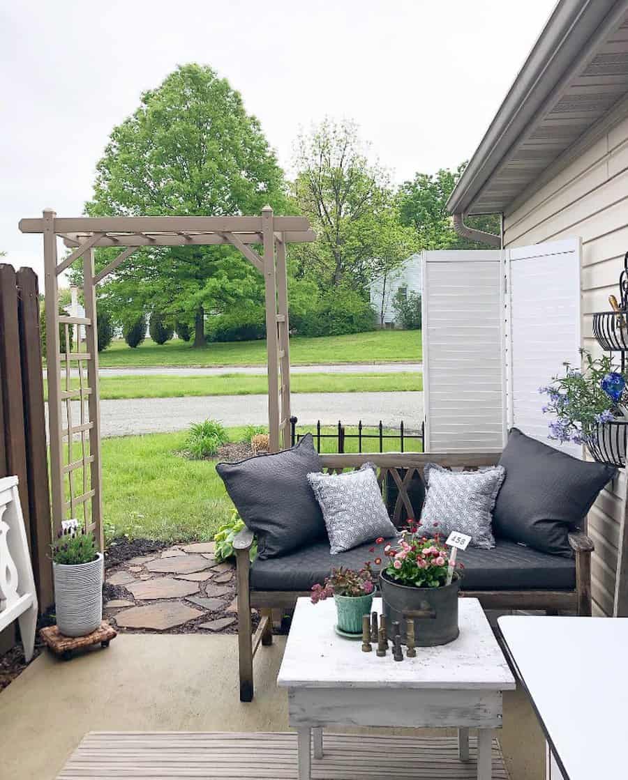 Cozy patio with a wooden bench, gray cushions, and decorative pillows. Potted plants on a white table, lattice archway, greenery beyond