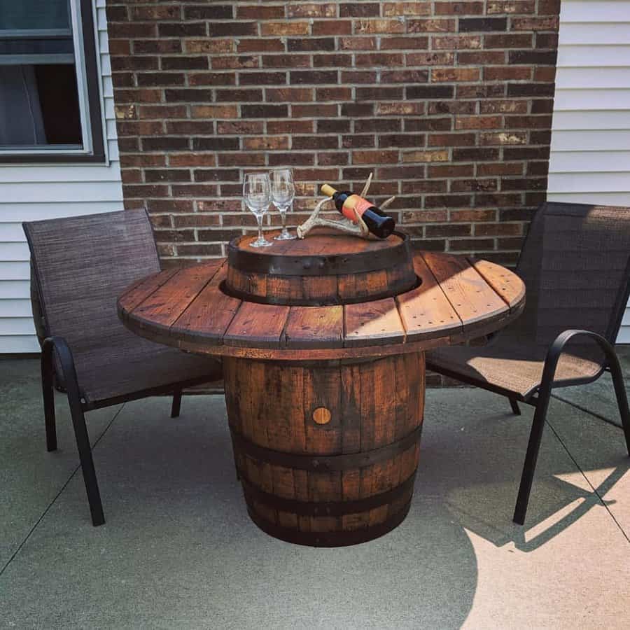 Wine barrel table with two glasses and a bottle, flanked by two chairs, against a brick and siding wall