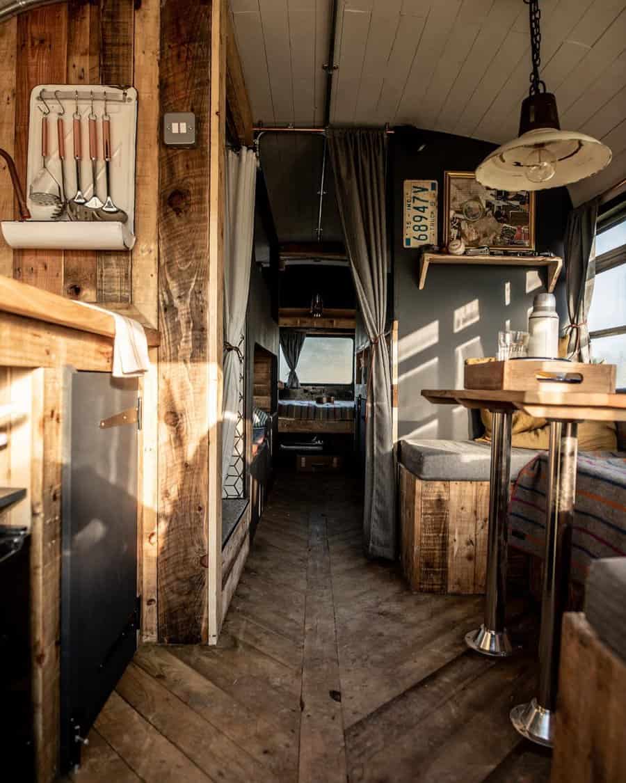 A rustic RV interior with a cozy and warm design featuring wooden walls, a herringbone floor, and a vintage light fixture, complemented by charming details like hanging kitchen tools and artwork.