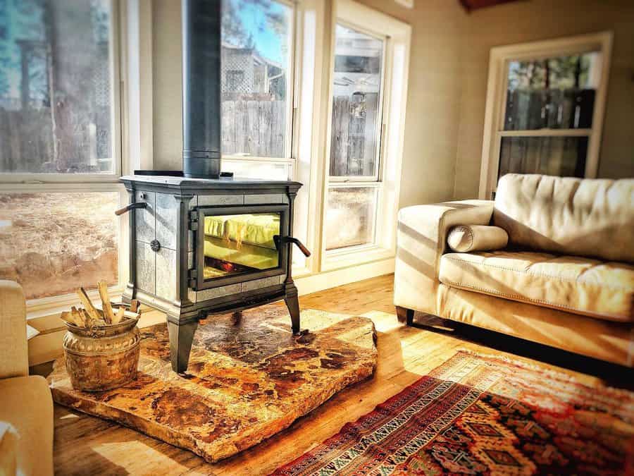 Freestanding wood stove on a rugged stone hearth, surrounded by large windows and warm furnishings for a cozy, sunlit retreat.