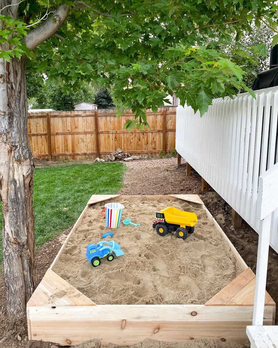 Children's sandbox with toy trucks, a striped bucket, and shovel, surrounded by grass, a wooden fence, and a tree providing shade