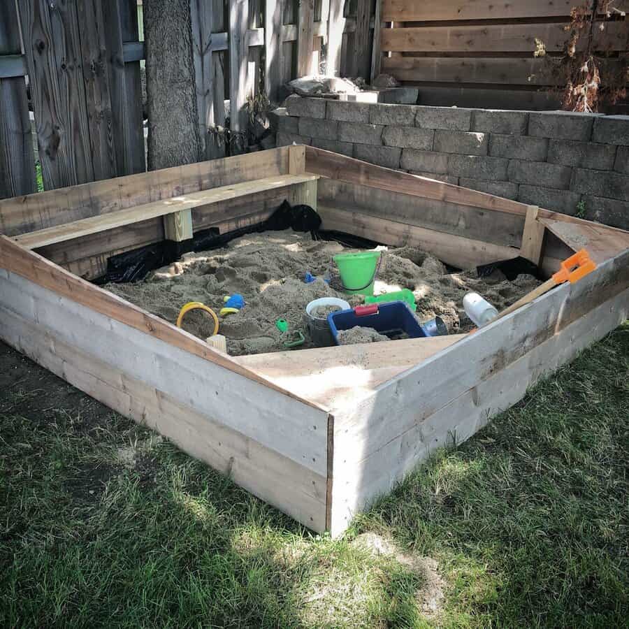 Square wooden sandbox filled with sand, toys, and buckets, set in a grassy backyard corner with a tree and wooden fence